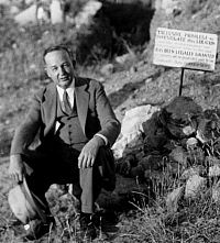 B&W photo of man dressed in a business suit seated beside posted sign at one of the ruins.
