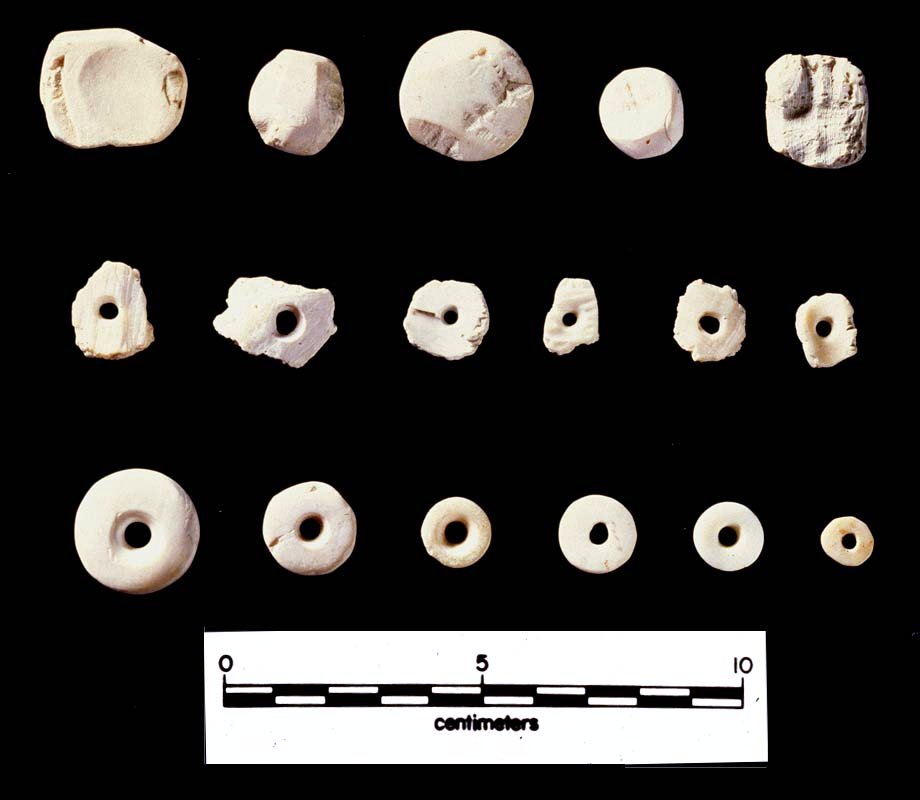 Shell-bead Making: The manufacture of round shell beads starts with "blanks," pieces of shell that have been broken or slightly rounded, such as the objects on the top row. On the middle row are roughed-out shell beads with drilled holes for suspension onto a necklace strand. Once the drilled beads were ground flat, they were tightly strung and the edges were ground and smoothed, strand-by-strand, creating uniform beads.