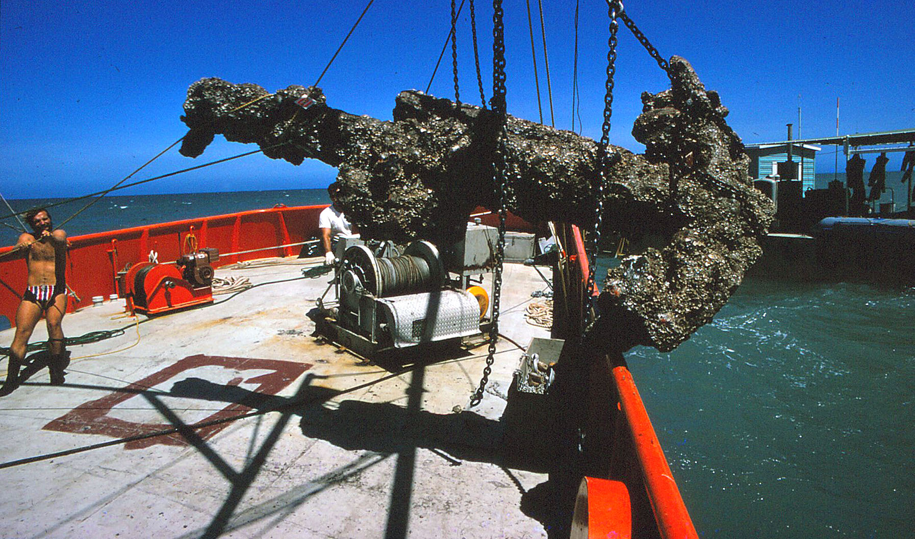 photo of man in striped speedo assisting in hoisting massive anchor onto deck of boat