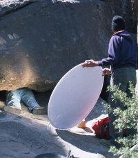 photo of Bob Mark of Rupestrian Cyberservices capturing a tight shot of rock art under a boulder while Evelyn Billo (left) and a crew member hold reflective panels to improve lighting