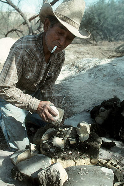 photo of man in a coyboy hat and plaid shirt working in a desert environment, smoking a cigarette and holding rock used to break up chunks of white material