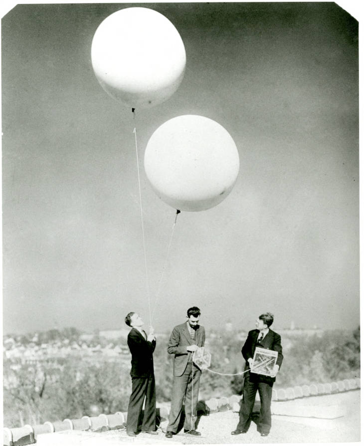 black and white photo of three men in suits below two weather balloons