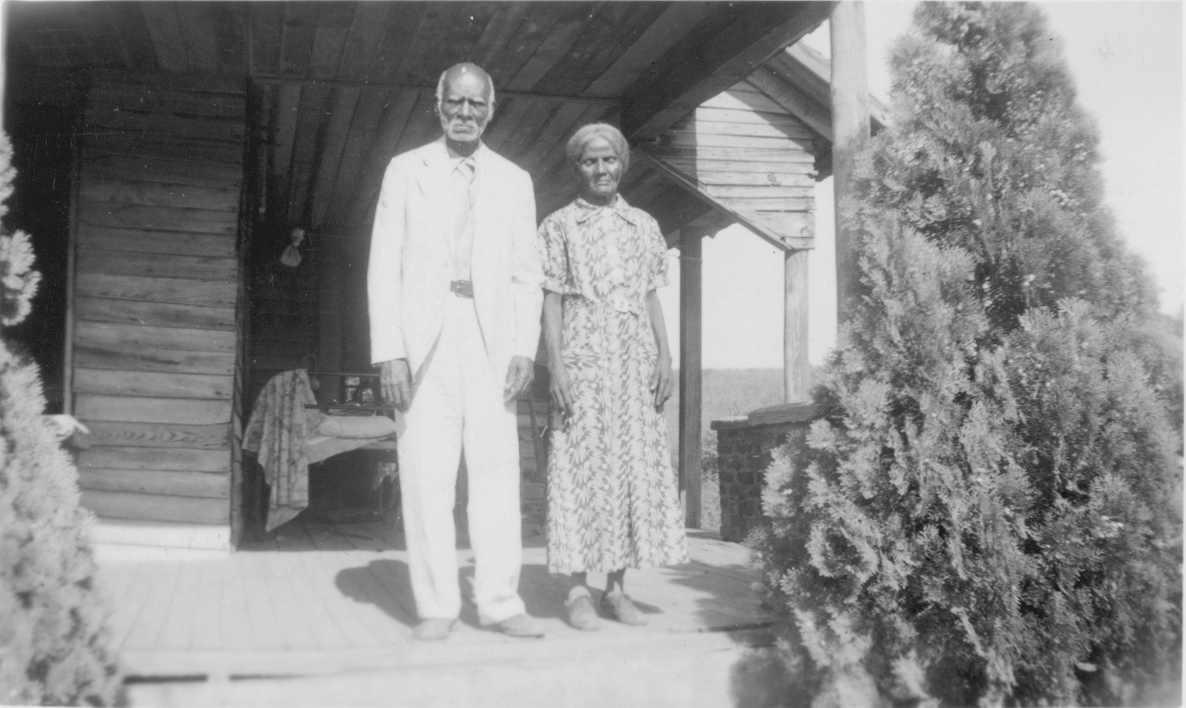 An older African American man and woman standing on a wood house porch. The man, on the right, has white hair and wears a white or light colored suit and the woman has her hair parted in the middle and pulled back. She wears a long pattered dress and her eyes are closed.