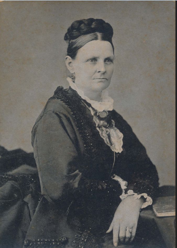 Black and white portrait of a Euro-American woman with her hair in an a braid wrapped around the top of her head, wearing an elaborate black dress, seated.