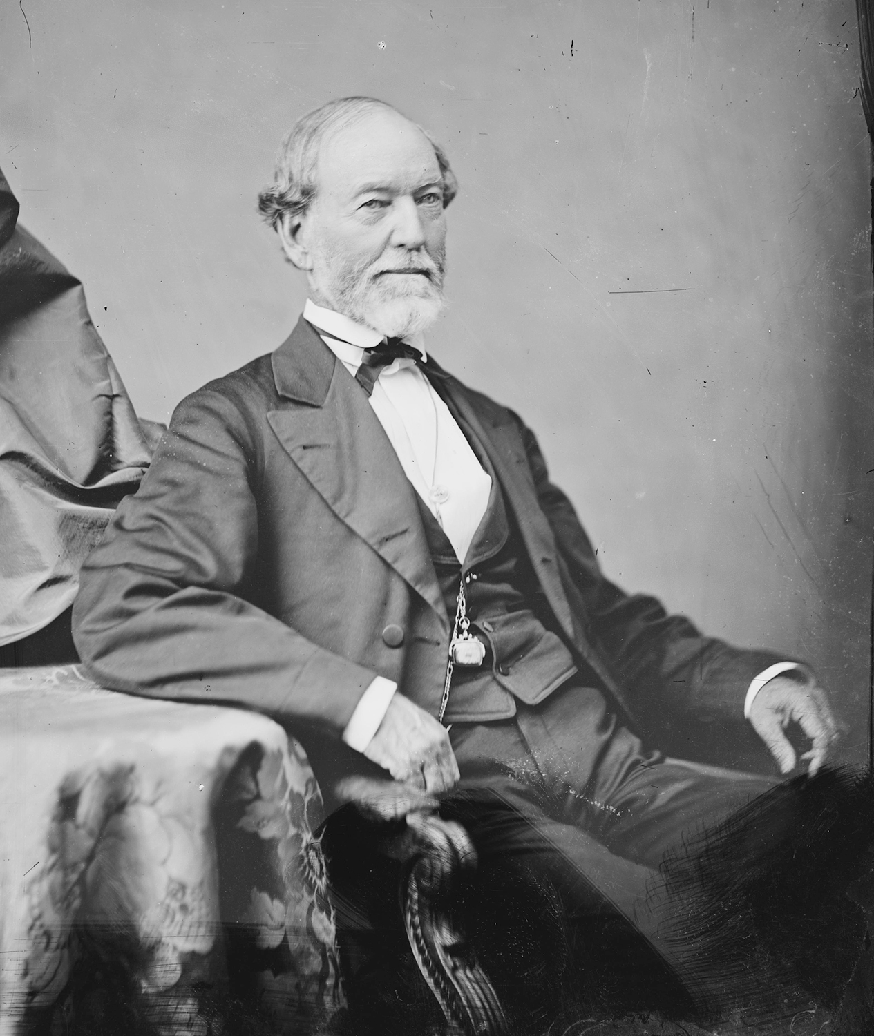 A black of white portrait of an older man wearing a suit, seated with his arm on a table on the left of the photograph.