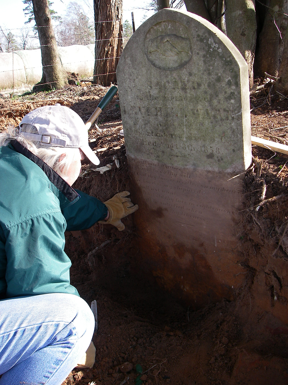 Photo of archeologist squating in excavation unit in front of headstone