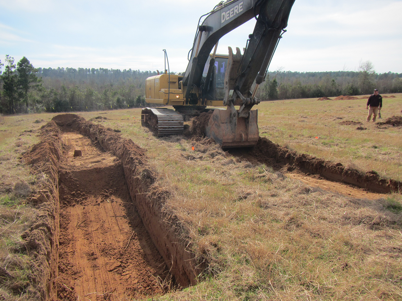 work shot photo; on left is open trench excavated into red sandy soil; on right a large trackhoe is digging a second trench