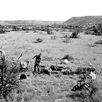 B&W photo of arid valley with man excavating a small test pit in foreground.