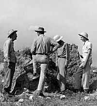 B&W photo showing four men dressed in field clothes deep in discussion in front of adobe wall. 